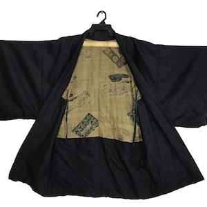 Puede incluir: Bata kimono negra con frente abierto, que revela un interior estampado. La bata est&aacute; colgada de una percha negra sobre un fondo blanco. La tela interior tiene un fondo beige con dise&ntilde;os geom&eacute;tricos y florales en verde oscuro y negro.