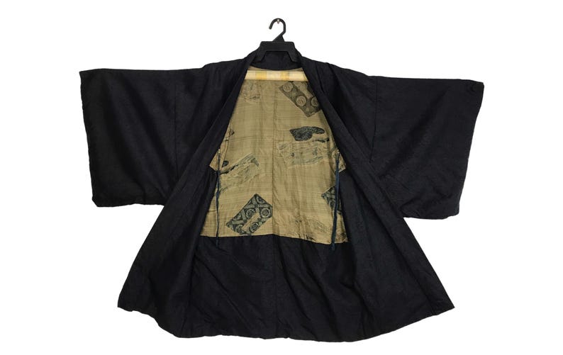 Puede incluir: Bata kimono negra con frente abierto, que revela un interior estampado. La bata est&aacute; colgada de una percha negra sobre un fondo blanco. La tela interior tiene un fondo beige con dise&ntilde;os geom&eacute;tricos y florales en verde oscuro y negro.