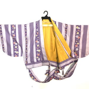 Puede incluir: Un kimono a rayas lavanda y blancas con detalles florales y un interior amarillo. El kimono está colgado de una percha negra, con las mangas extendidas. Los detalles florales incluyen flores rosas, amarillas y blancas.