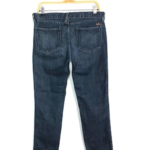 May include: Dark wash denim jeans with two back pockets and a small orange logo on the back. The jeans are hanging on a wooden hanger against a white background. The jeans are a classic, straight-leg style.