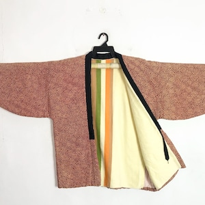 Puede incluir: Chaqueta vintage estilo kimono con un estampado de lunares rojos y marrones. La chaqueta tiene un forro color crema y un panel a rayas con colores verde, naranja y crema. Los bordes están ribeteados con tela negra.