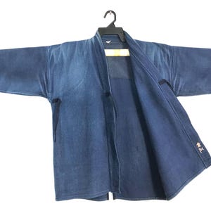 May include: A blue denim jacket with a V-neck design, hanging on a black hanger. The jacket has long sleeves and a textured appearance. The jacket is open in the front and has a small design on the lower right side.