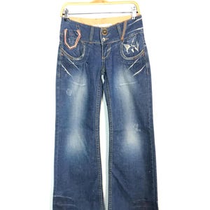 Op de afbeelding: Blauwe denim jeans met een wijde pijp. De jeans hebben decoratieve stiksels, distressed details en een knoopsluiting. De zakken hebben een uniek ontwerp met roze accenten. De jeans hangen aan een houten hanger.