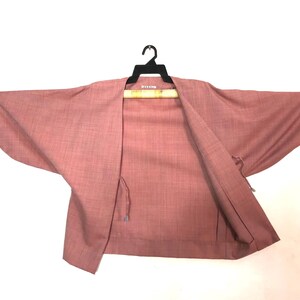 Puede incluir: Chaqueta estilo kimono rosa polvoriento con mangas anchas, colgada de una percha negra. La parte delantera abierta revela el interior de la chaqueta y se ve un detalle de lazo. La tela tiene una textura sutil.