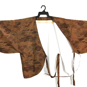 May include: A brown kimono jacket featuring a detailed landscape print with houses, trees, and rolling hills. The garment has wide sleeves, an open front, and matching fabric ties attached to the side. The interior is lined with a plain white fabric.