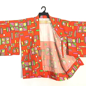 May include: A bright orange kimono featuring a repeating geometric pattern in shades of lime green, hot pink, and white. The garment has wide sleeves, an open front, and a white interior lining, displayed on a black plastic clothes hanger.