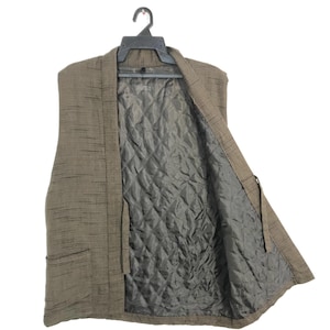 May include: A brown, sleeveless vest with a textured exterior and a quilted, gray interior. The vest is hanging on a black hanger against a white background. It has two front pockets and a tie closure.
