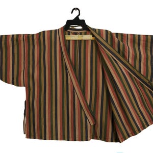 May include: A striped kimono-style jacket in shades of brown, green, and red hangs on a black hanger. The open front reveals the interior lining. The vertical stripes create a visually appealing pattern.