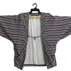 May include: A gray kimono-style jacket with horizontal stripes in burgundy and black. The jacket has wide sleeves and a white interior. It is hanging on a black hanger.