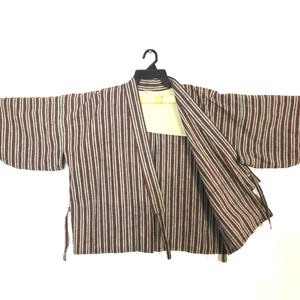 May include: A striped kimono-style jacket with vertical burgundy, beige, and white stripes. The jacket has wide sleeves and is open at the front, revealing a cream-coloured lining. It hangs from a black hanger.