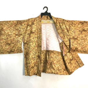 May include: A vintage kimono with a yellow and brown floral pattern, hanging on a black hanger. The kimono's interior lining is a light pink color. The sleeves are wide and the fabric appears to be lightweight.