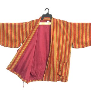 May include: A striped orange and yellow kimono-style jacket with a red interior. The jacket is hanging on a black hanger against a white background. The sleeves are wide and the jacket has two pockets.
