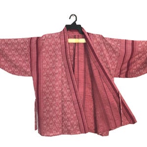 May include: A pink kimono-style jacket featuring a repeating geometric pattern on the sleeves and front panels, contrasted by solid vertical stripes. The garment has wide sleeves with gathered cuffs, an open front, and a tie closure at the side.
