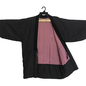 Puede incluir: Un kimono negro con un sutil estampado de puntos y un interior rosa contrastante. El kimono está abierto, revelando el forro y la forma de la prenda. Cuelga de una percha negra.