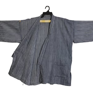 Puede incluir: Chaqueta estilo kimono a rayas azules y blancas colgada de una percha negra. La chaqueta tiene mangas cortas y dos bolsillos delanteros. La tela tiene un aspecto texturizado.
