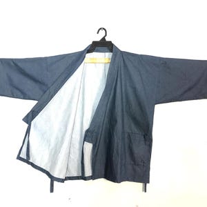 May include: A blue kimono-style jacket with long sleeves, hanging on a black hanger. The jacket has a white interior and two front pockets. The jacket is made of a lightweight fabric.
