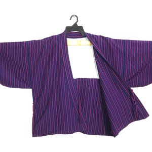 May include: A dark purple kimono-style jacket featuring thin, vertical magenta stripes throughout the fabric. The garment has wide, loose sleeves, an open front design, and a white inner lining visible at the collar area, displayed on a black hanger.