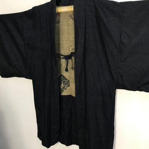 Puede incluir: Chaqueta negra estilo kimono con un patr&oacute;n texturizado. La chaqueta est&aacute; abierta, revelando un forro beige con un dise&ntilde;o decorativo. Cuelga de una percha negra sobre un fondo blanco. La chaqueta tiene mangas anchas y un cierre de lazo.