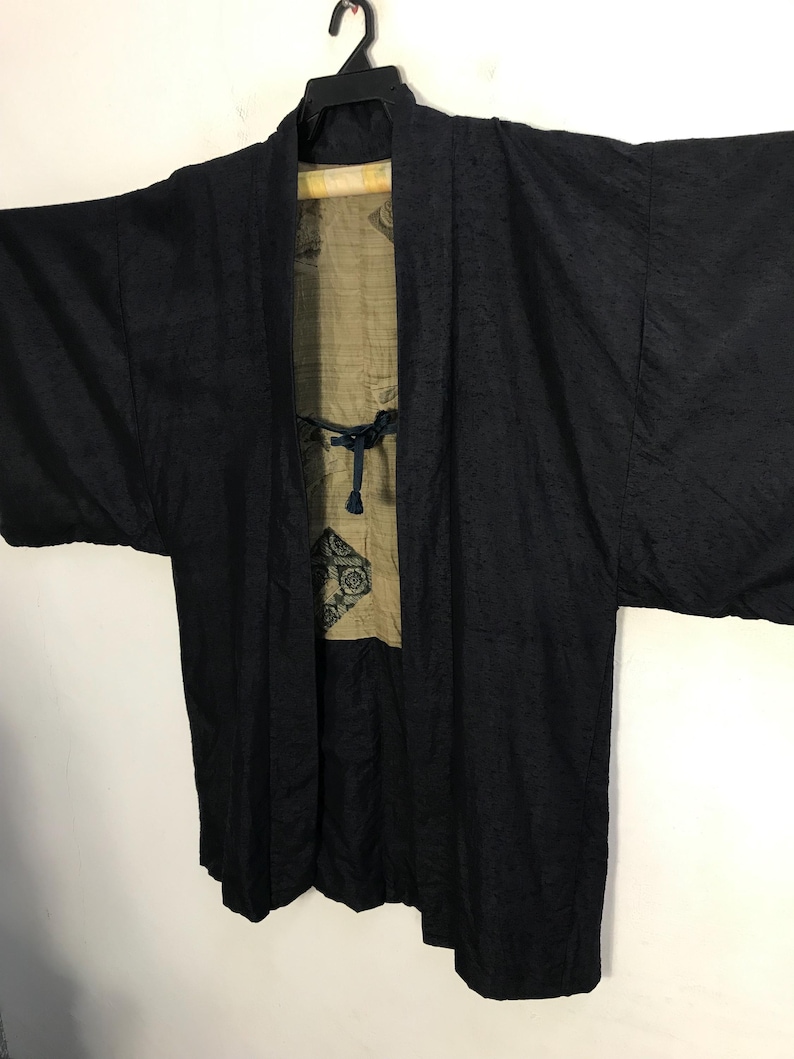 Puede incluir: Chaqueta negra estilo kimono con un patr&oacute;n texturizado. La chaqueta est&aacute; abierta, revelando un forro beige con un dise&ntilde;o decorativo. Cuelga de una percha negra sobre un fondo blanco. La chaqueta tiene mangas anchas y un cierre de lazo.
