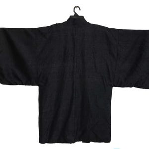 Puede incluir: Una bata negra estilo kimono con mangas anchas, colgada de una percha negra. La bata tiene un patr&oacute;n texturizado y un cuello alto. La prenda se muestra sobre un fondo blanco.
