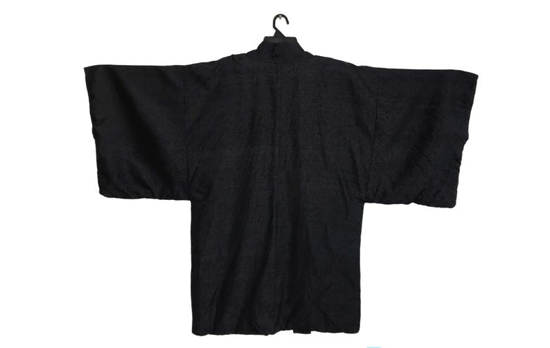 Puede incluir: Una bata negra estilo kimono con mangas anchas, colgada de una percha negra. La bata tiene un patr&oacute;n texturizado y un cuello alto. La prenda se muestra sobre un fondo blanco.