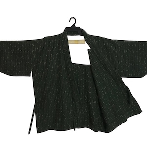 Puede incluir: Una chaqueta verde oscuro estilo kimono con un patrón de rayas verticales. La chaqueta tiene mangas anchas y está colgada de una percha negra. La tela parece tener textura y la chaqueta está abierta por delante.