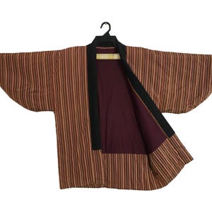 Puede incluir: Una chaqueta estilo kimono a rayas marrones y beige con cuello negro y forro burdeos, colgada de una percha negra. La chaqueta está abierta, mostrando el forro interior.