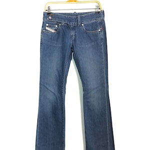 Könnte beinhalten: Blaue Jeans aus Denim mit leicht ausgestelltem Bein, hängen an einem goldfarbenen Kleiderbügel. Die Jeans haben ein klassisches Fünf-Taschen-Design, einen Knopf- und Reißverschluss und ein kleines Logo auf der Vordertasche.