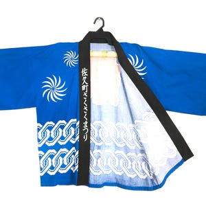 May include: A bright blue happi coat featuring a black collar with white Japanese text, white spiral patterns on the upper section, and a white chain-link design along the bottom hem. The garment is displayed on a black plastic hanger.