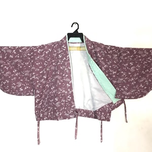 May include: A purple floral patterned kimono with wide sleeves, hanging on a black hanger. The inside lining is white and light green. The kimono has tie closures at the waist.