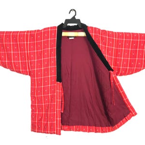May include: A red quilted jacket with a grid pattern and black trim, hanging on a black hanger. The jacket's interior is a solid burgundy color. The sleeves are wide and the jacket is open in the front.