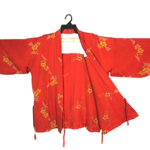 May include: A bright red haori jacket featuring a repeating pattern of yellow floral clusters and light gray geometric shapes. The garment has wide, open sleeves, a front opening, and thin red fabric ties attached to the lower side seams.