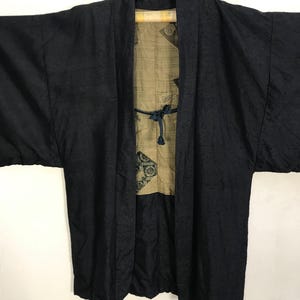 Puede incluir: Chaqueta negra estilo kimono con exterior texturizado e interior estampado. La chaqueta est&aacute; colgada de una percha negra. El forro presenta un fondo beige con dise&ntilde;os florales en verde oscuro y negro. Una corbata azul asegura la chaqueta.