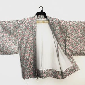 May include: A short kimono-style jacket featuring a repeating floral pattern in shades of pink and grey on a light background. The garment has wide, rectangular sleeves, an open front, and a solid white lining visible on the interior panels.