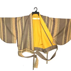 May include: A kimono-style robe with a yellow interior and a patterned exterior in shades of beige, brown, and olive green. The robe is hanging on a black hanger against a white background. The robe has long, wide sleeves and a tie closure.