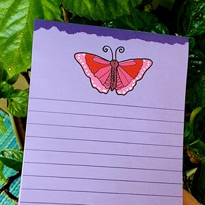 May include: A lined lavender notepad with a torn edge design. A pink and red butterfly illustration is at the top. The notepad is held against a background of green leaves.