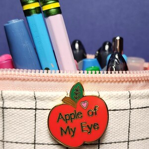 May include: A pencil case with a red apple-shaped pin that reads "Apple of My Eye." The case contains pencils and pens in various colors, including blue, pink, and green. The case has a pink zipper and a white grid pattern.