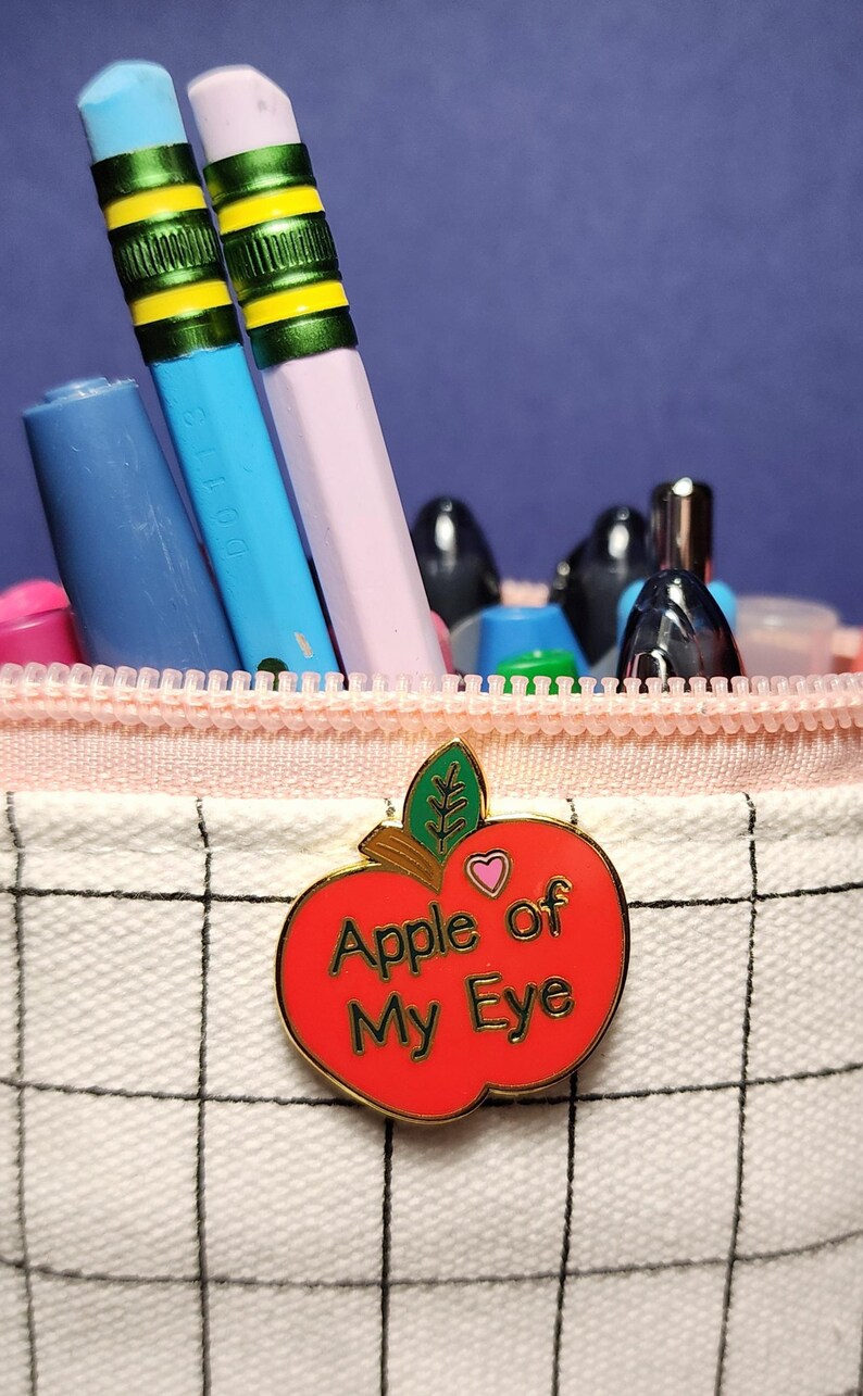 May include: A pencil case with a red apple-shaped pin that reads "Apple of My Eye." The case contains pencils and pens in various colors, including blue, pink, and green. The case has a pink zipper and a white grid pattern.