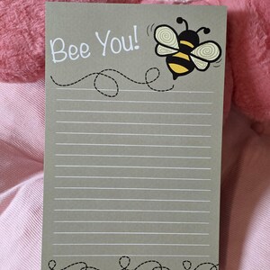 May include: A lined notepad with the words "Bee You!" in white, featuring a cartoon bee with yellow and black stripes. The notepad has a gray background with a dotted line design.