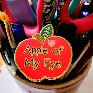 May include: A red enamel apple-shaped pin with gold trim and the words "Apple of My Eye." The pin is surrounded by pens, scissors, and a paintbrush in a decorative ceramic holder.