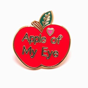 May include: A red enamel apple-shaped pin with gold trim and the words "Apple of My Eye" in gold lettering. The pin features a green leaf and a pink heart. A charming accessory.