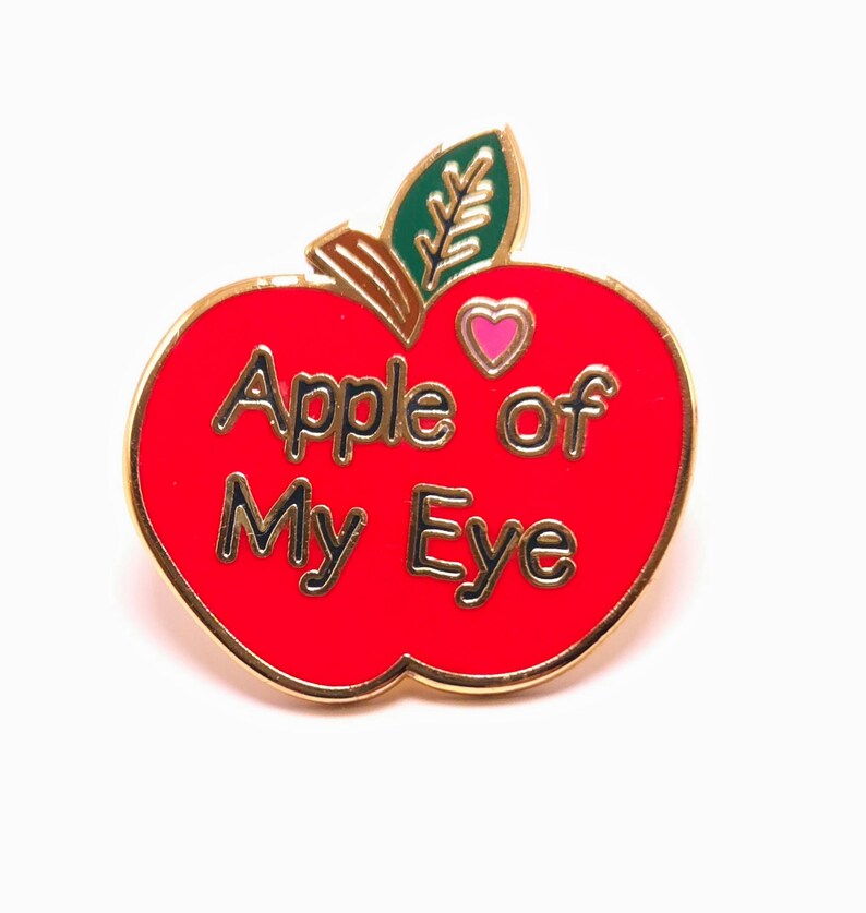 May include: A red enamel apple-shaped pin with gold trim and the words "Apple of My Eye" in gold lettering. The pin features a green leaf and a pink heart. A charming accessory.