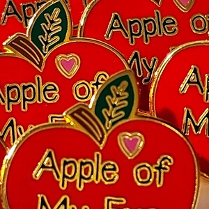 May include: Close-up of several red apple-shaped pins with gold trim. Each pin features the phrase "Apple of My Eye" in gold lettering, a pink heart, and a green leaf. The pins are likely decorative accessories.