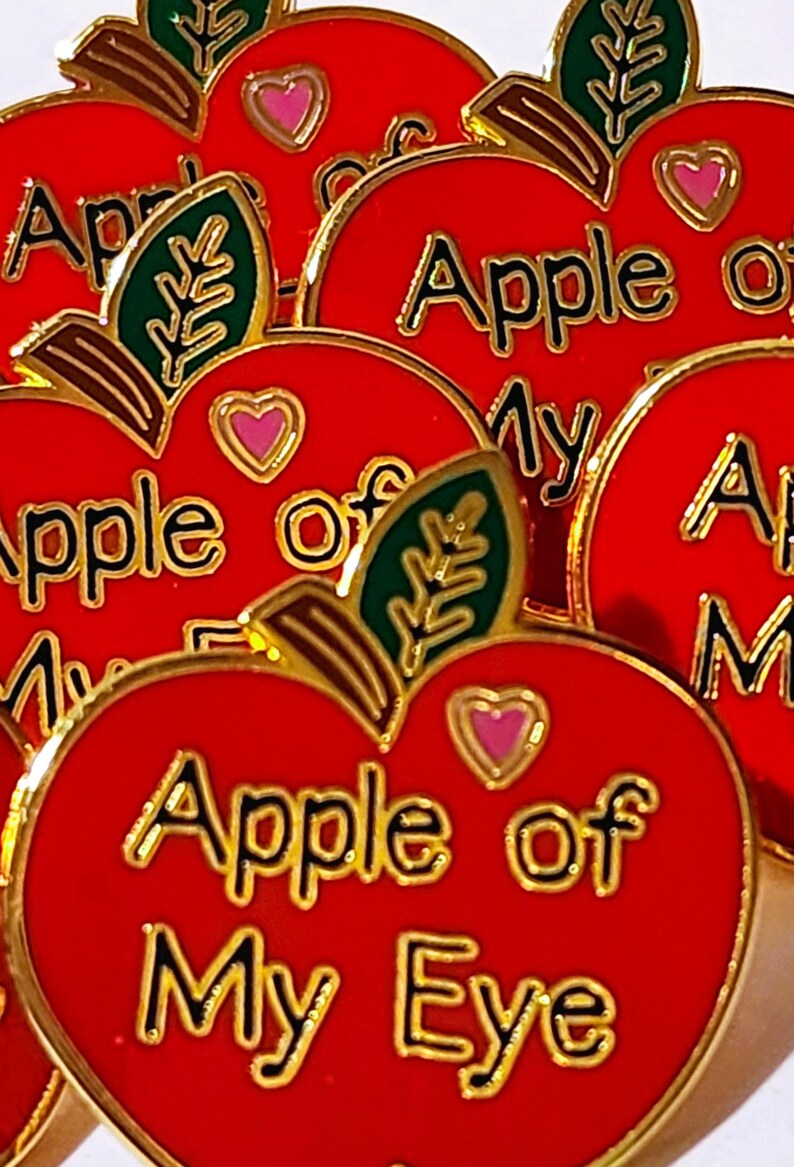 May include: Close-up of several red apple-shaped pins with gold trim. Each pin features the phrase "Apple of My Eye" in gold lettering, a pink heart, and a green leaf. The pins are likely decorative accessories.