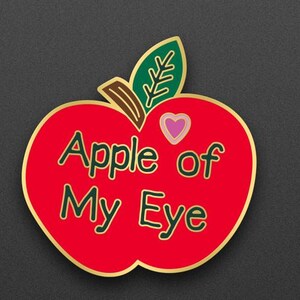May include: A red apple-shaped pin with the text "Apple of My Eye" in green lettering. The pin has a gold border, a green leaf, and a pink heart. The design is on a dark gray background.