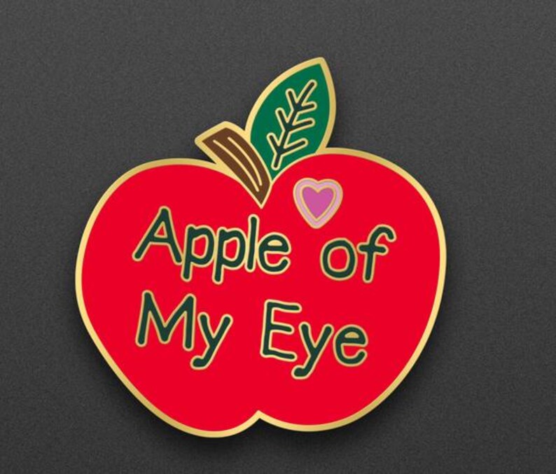 May include: A red apple-shaped pin with the text "Apple of My Eye" in green lettering. The pin has a gold border, a green leaf, and a pink heart. The design is on a dark gray background.