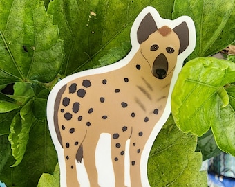 May include: A sticker featuring a cartoon hyena with a tan body, black spots, and dark brown ears. The hyena is standing on a white background, set against a backdrop of green leaves.