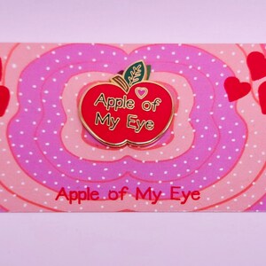 May include: A red enamel pin in the shape of an apple with the text "Apple of My Eye" in gold lettering. The pin is on a pink and white patterned card with the same text. Red heart shapes are scattered around the pin.
