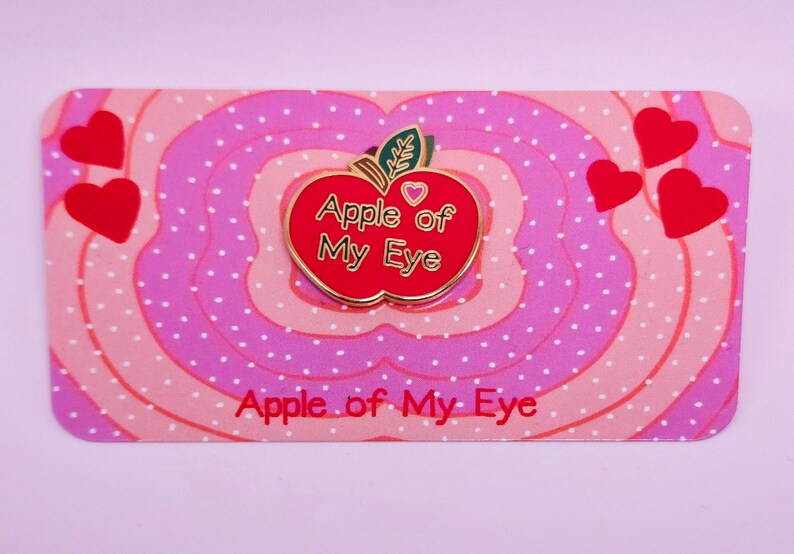 May include: A red enamel pin in the shape of an apple with the text "Apple of My Eye" in gold lettering. The pin is on a pink and white patterned card with the same text. Red heart shapes are scattered around the pin.