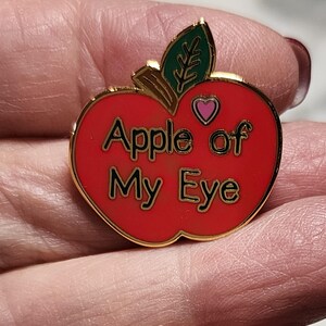 1 inch enamel pin in the shape of an apple.  Red with a small pink heart at top for Valentines.  Green leaf and Brown Stem. Shiny gold background and back. Smooth.  Hard Enamel.  Text on apple reads, "Apple of My eye."